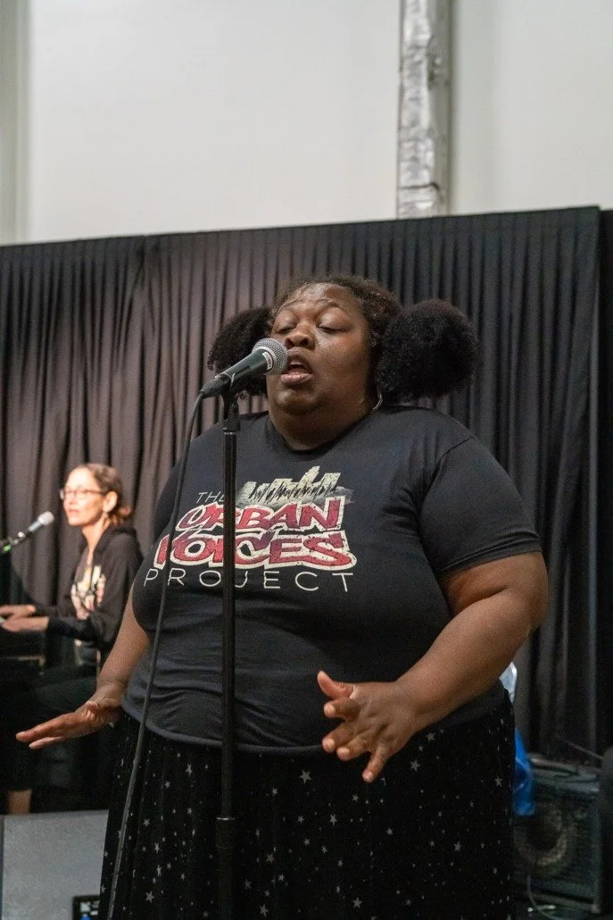 A woman singing into a microphone with her eyes closed, wearing a Black Lives Matter T-shirt, with a woman playing a keyboard in the background.