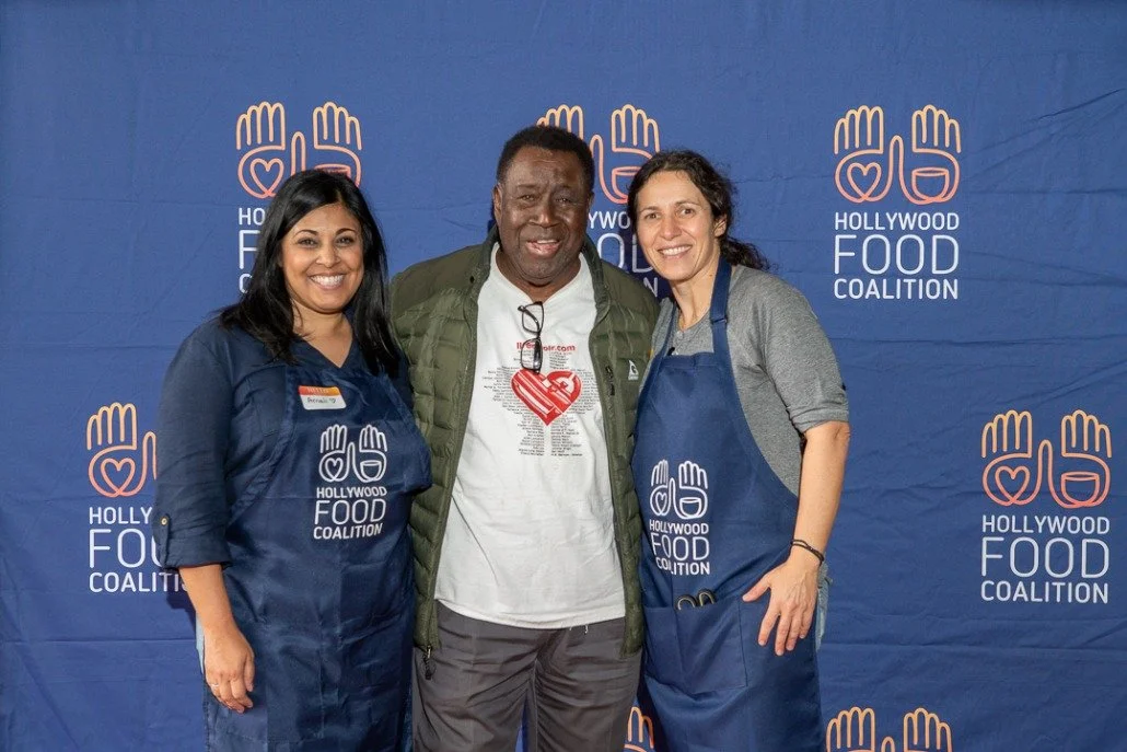 Three people standing together at a Hollywood Food Coalition event, smiling for the camera, two women wearing blue aprons with the coalition's logo, man in the middle wearing a white t-shirt and green jacket, with a background displaying the Hollywoo