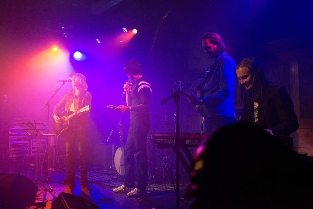 Band performing on stage with colorful lighting, featuring a guitarist, singer, and two keyboard players.