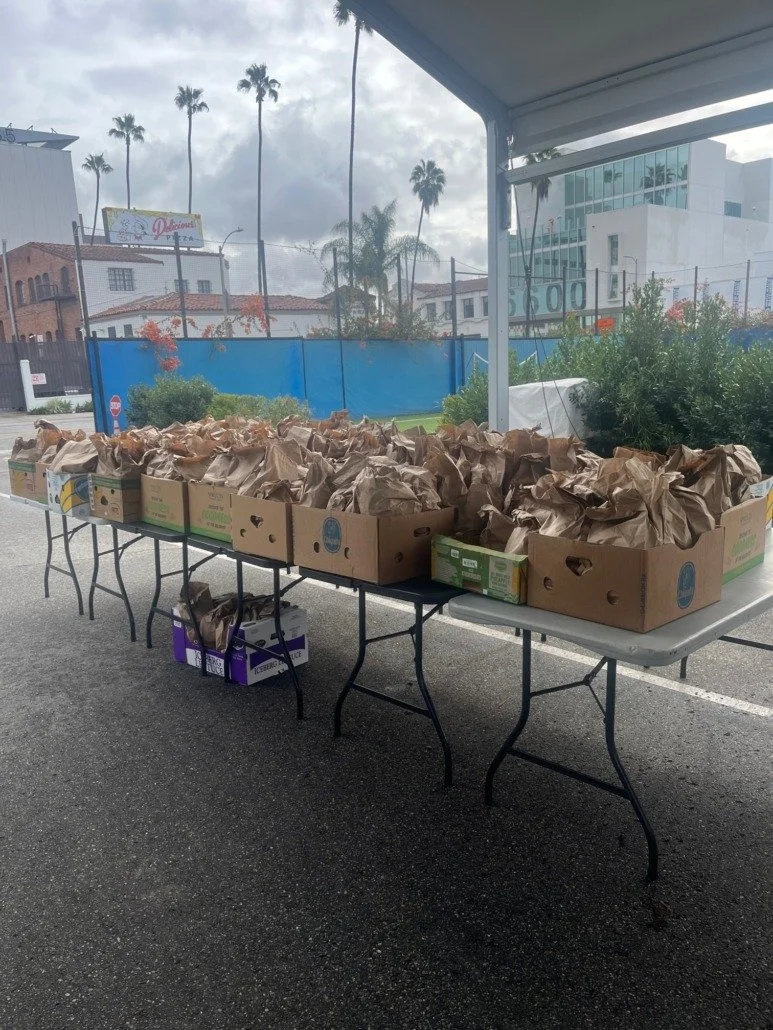Tables set up outdoors with boxes of produce and paper bags, with trees and buildings in the background.