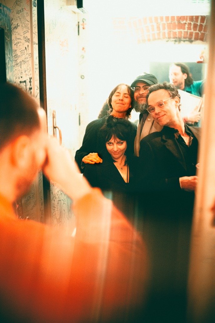 Group of five people taking a selfie in a mirror, smiling, with graffiti-covered wall and brick wall in background.