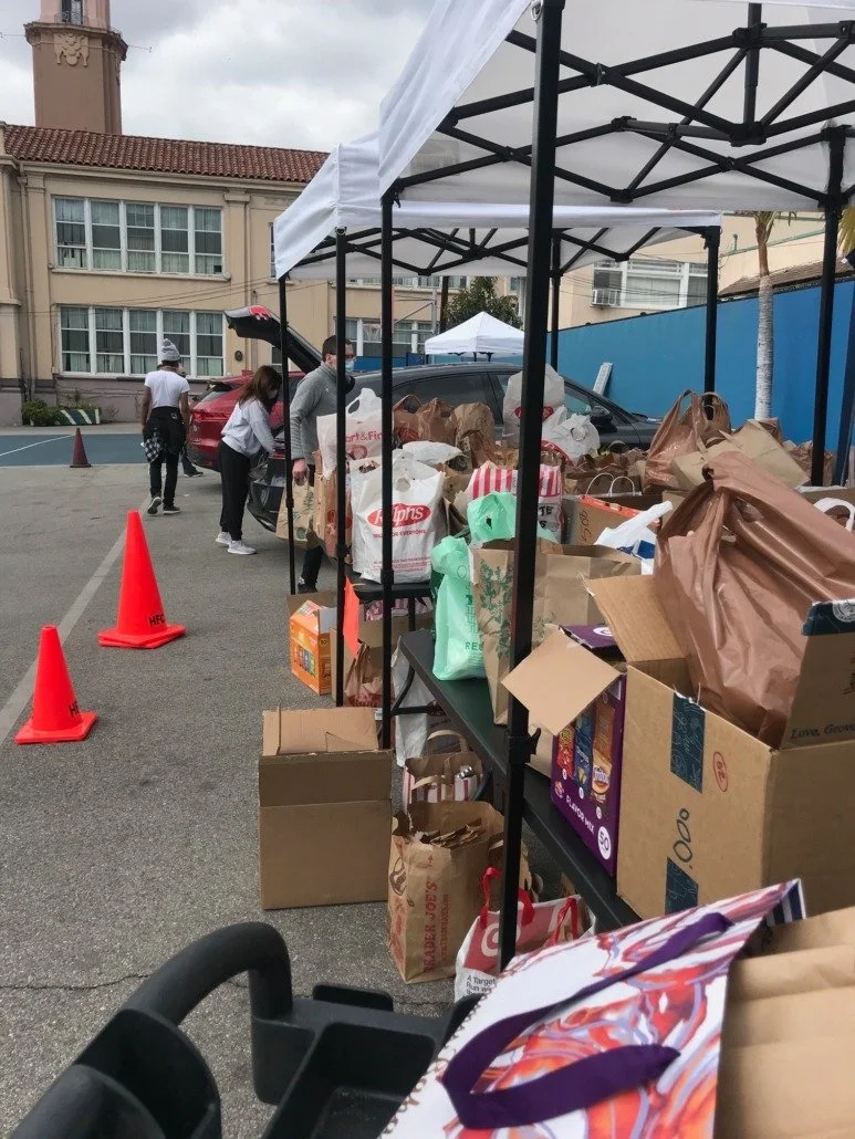 Pop-up outdoor donation or giveaway event with tables filled with bags and boxes of items, orange traffic cones marking the area, and people browsing or collecting items, set against a background of buildings and cloudy sky.
