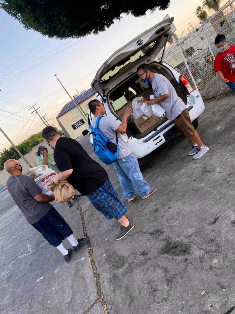 People loading supplies into the back of a white SUV in an outdoor parking lot, with others standing nearby and a chain-link fence in the background.
