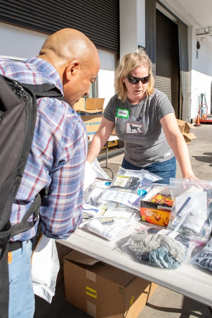 A woman with sunglasses and a gray t-shirt showing a dog and a man with a backpack examining various items on a table outside a building.