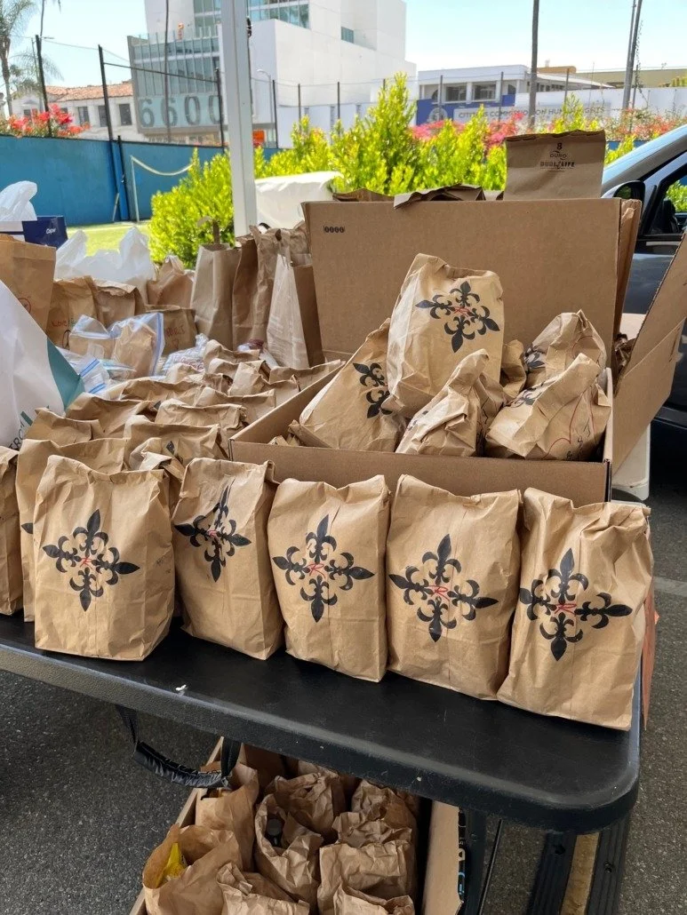 Multiple paper bags with a fleur-de-lis pattern on a black table outdoors, with a cardboard box and a car visible in the background.