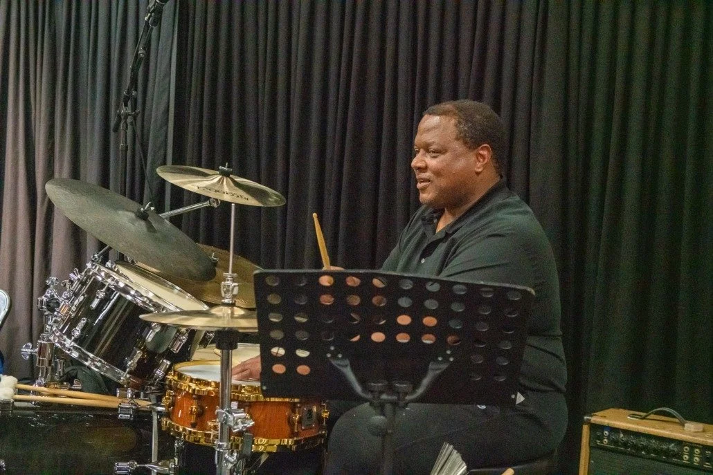 A man playing the drums on stage with black curtains in the background.