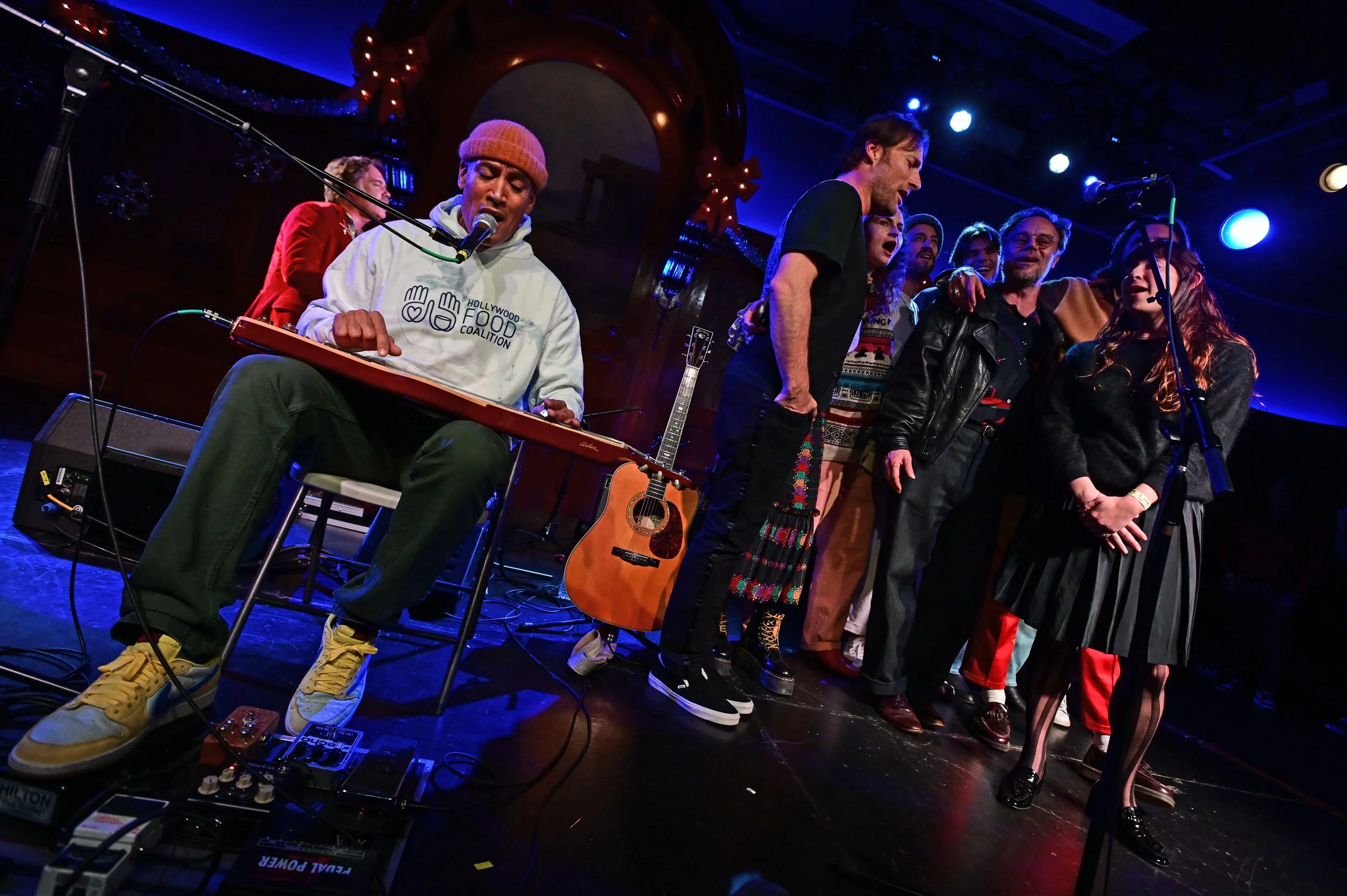 A group of people singing together on stage, with one person playing a pedal steel guitar, in a dimly lit venue decorated for Christmas with red ribbons and lights.