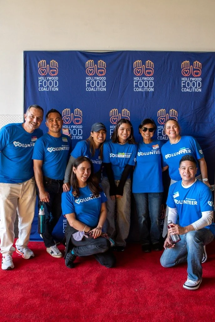 A group of seven volunteers wearing blue shirts with the word 'Volunteer' printed on them, standing and kneeling on a red carpet in front of a blue backdrop with the 'Hollywood Food Coalition' logo. They are smiling at the camera.