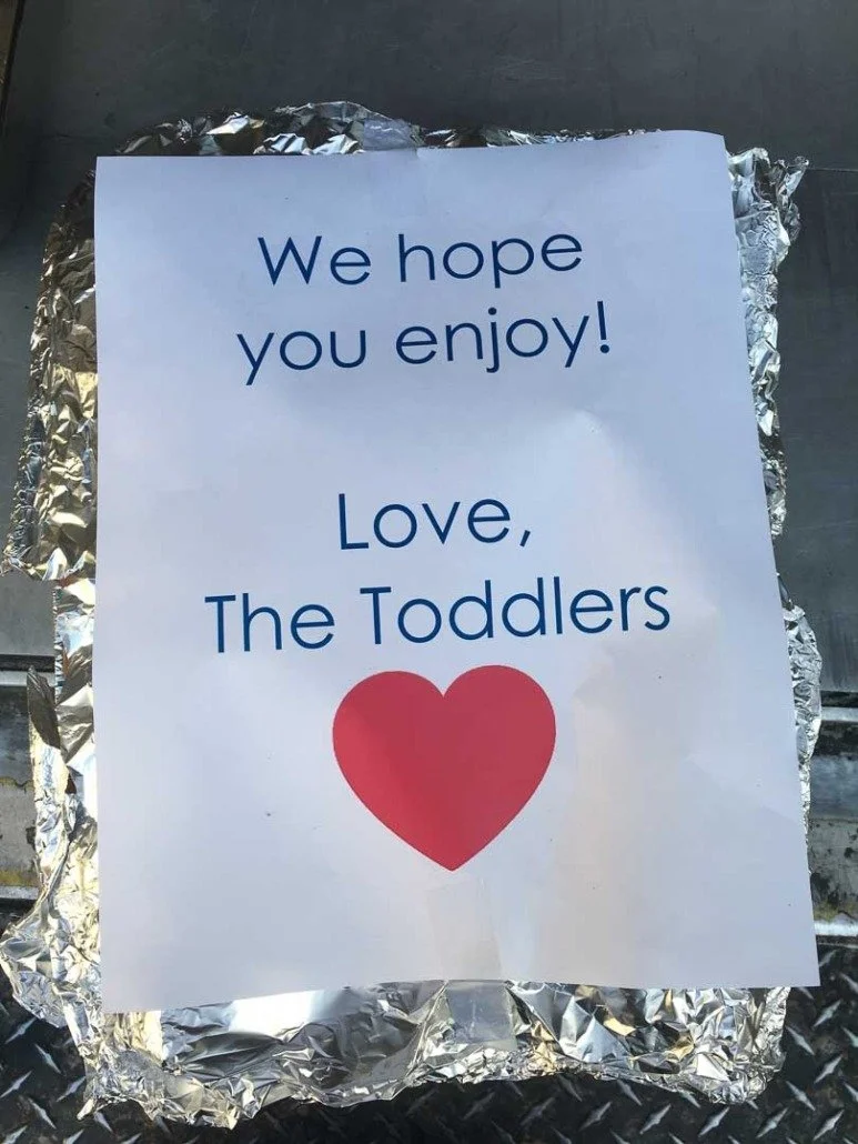 A handwritten note on white paper with blue text and a red heart, surrounded by crumpled aluminum foil. The note reads, "We hope you enjoy! Love, The Toddlers."