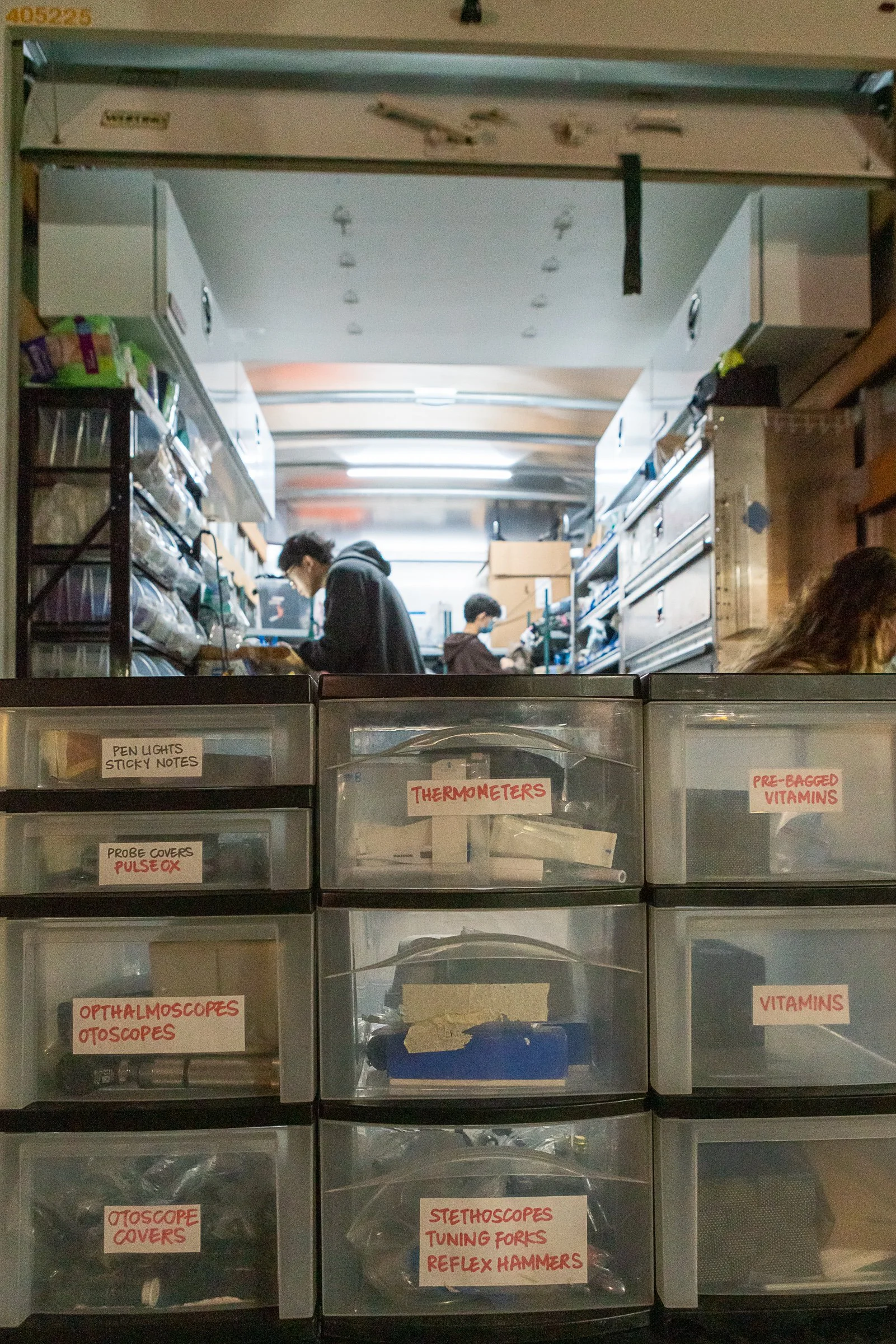 Medical supply storage with labeled drawers including thermometers, pens, sticky notes, pre-bagged vitamins, probe covers, pulse ox, ophthalmoscopes, otoscopes, tuning forks, reflex hammers, stethoscopes, and flashlights, in a medical setting with tw