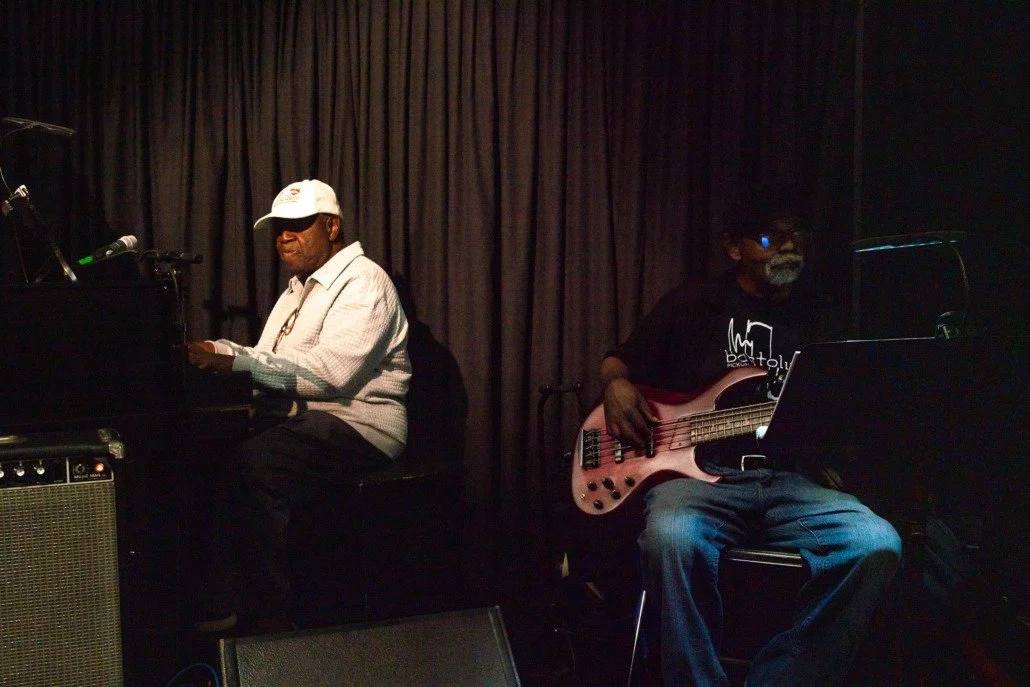 Two musicians performing on stage with dark curtains behind them, one playing the piano and the other playing a bass guitar, both men wearing casual clothing and sunglasses.