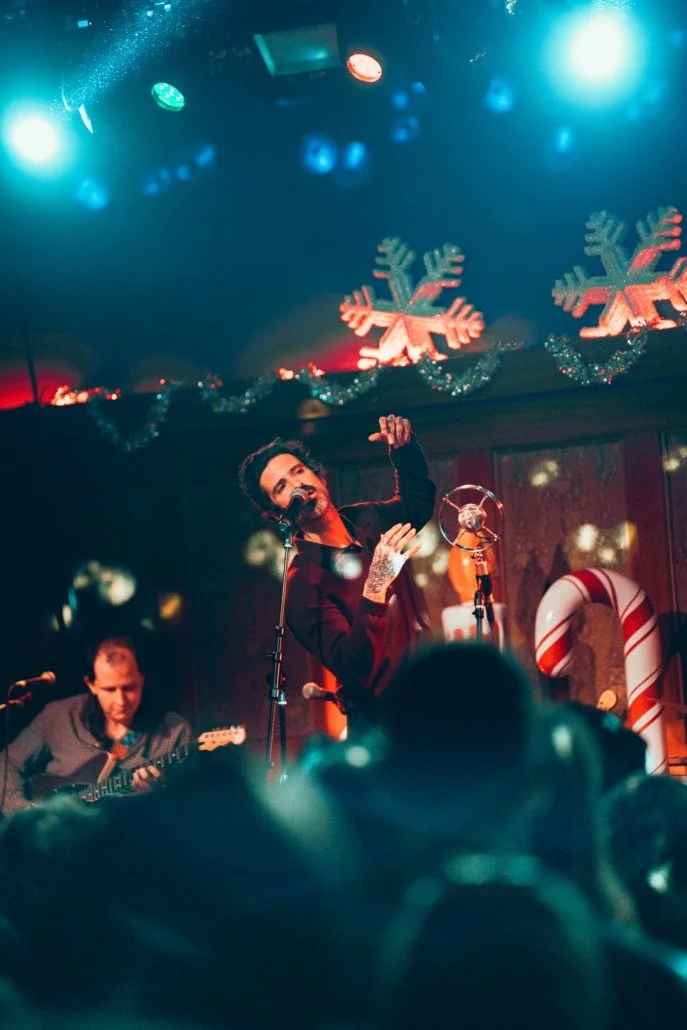 A singer with dark hair, wearing a black outfit, performing on stage with a microphone, during a Christmas-themed concert with snowflake and candy cane decorations.