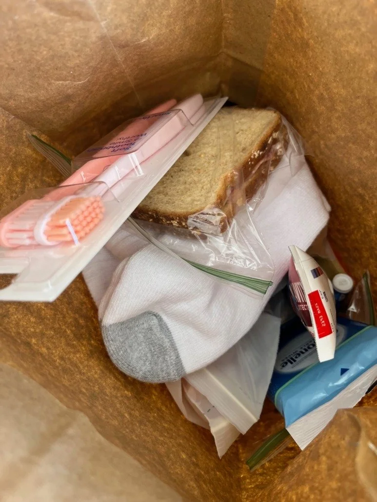 Inside a brown paper bag containing a sandwich in plastic wrap, a sock, toothbrush in plastic packaging, a tube of toothpaste, and other miscellaneous items.