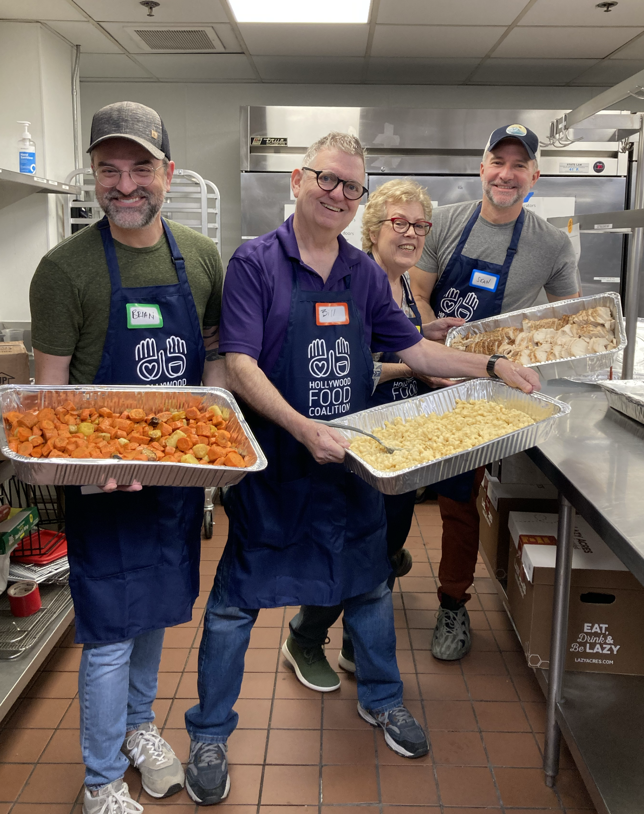 Four smiling people in aprons standing in a kitchen holding trays of food, with the group labeled with name tags: Brian, Bill, and Sean. The trays contain cooked vegetables, rice, and sliced cooked chicken.