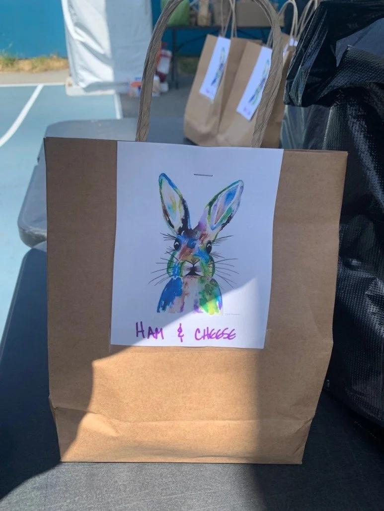 A brown paper bag with a colorful watercolor illustration of a rabbit and the words "Ham & Cheese" written in purple underneath. In the background, other paper bags with similar labels and a black plastic bag are visible, set outdoors on a picnic tab