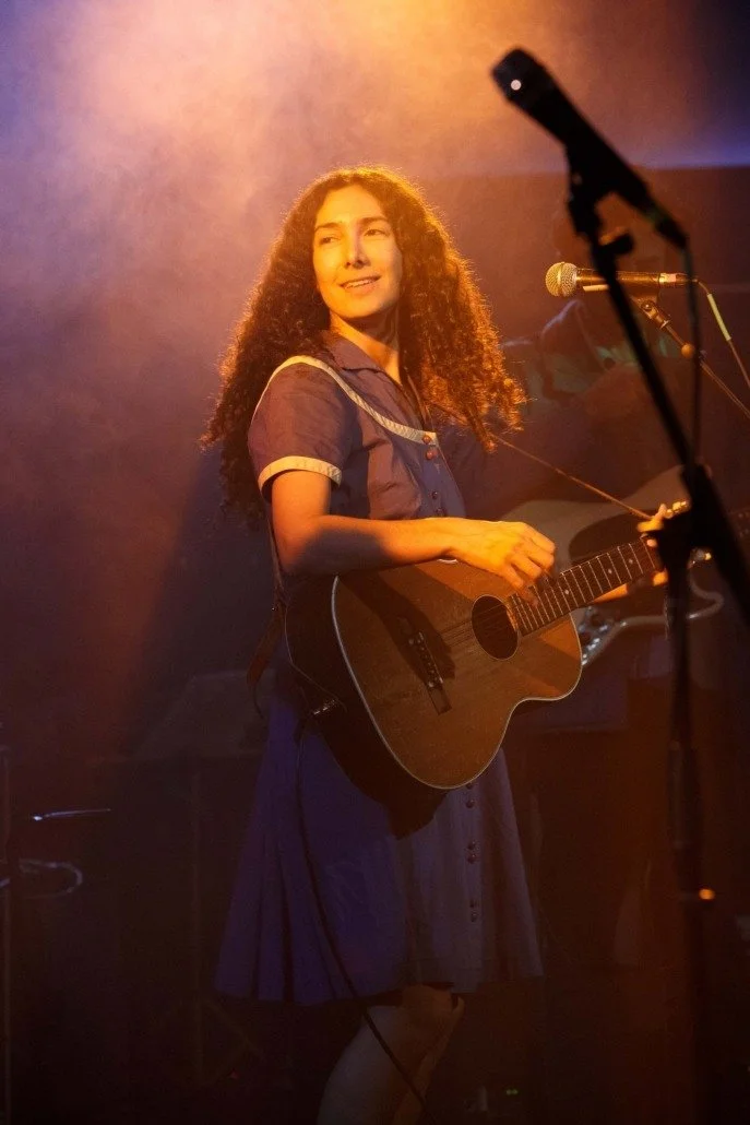 A woman with long curly hair plays an acoustic guitar on stage, smiling amidst warm stage lighting.