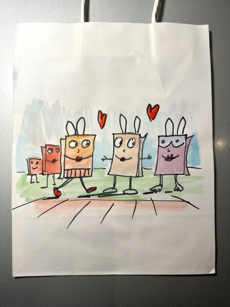 Drawing of four anthropomorphic paper bag characters with bunny ears, holding hands and walking outdoors with a grassy background. Two small characters in the background, one smiling and the other peeking out from behind.