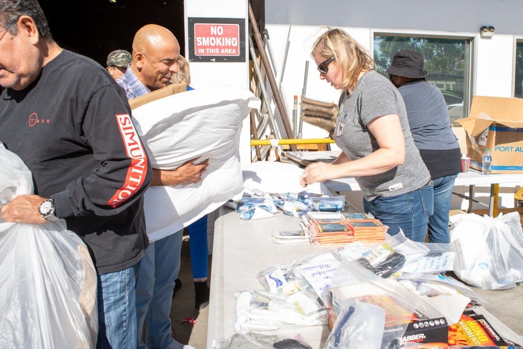 People volunteering at a food or supply drive, distributing items like water bottles, notebooks, and snacks at an outdoor setup with tables and supplies.