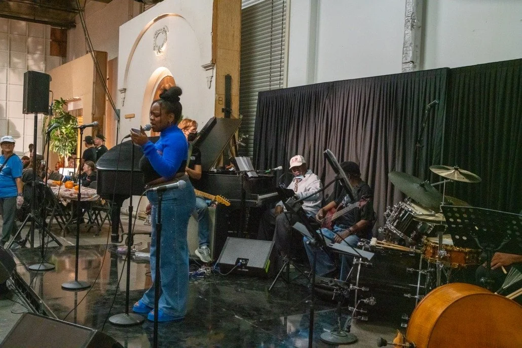 A woman in a blue top and jeans singing into a microphone on a stage, accompanied by band members playing piano, guitar, drums, and other instruments at an indoor event.