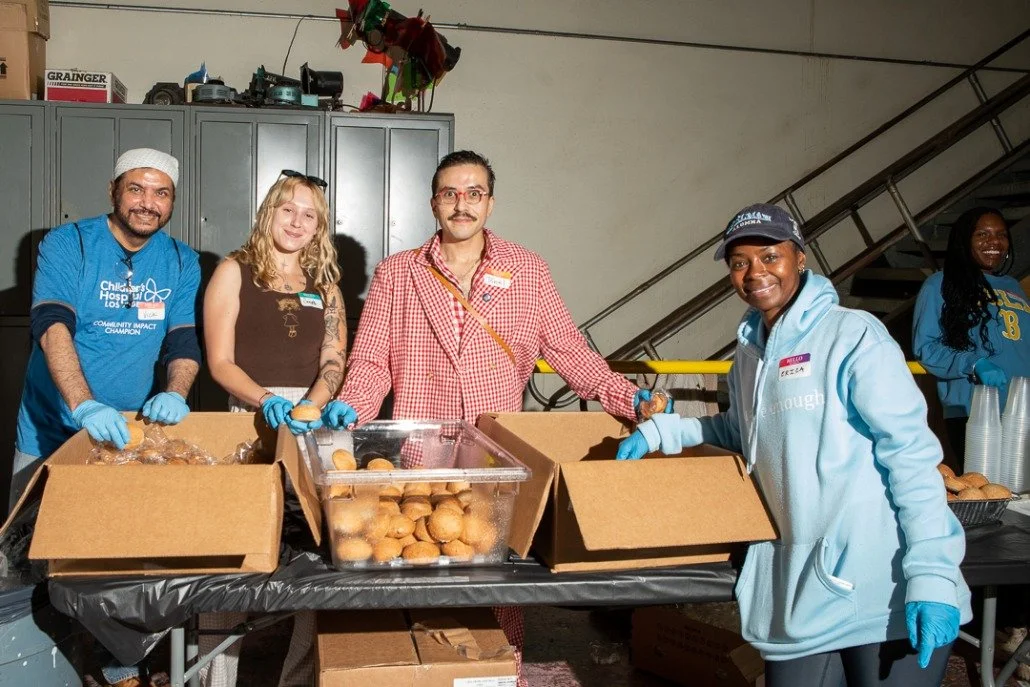 Group of five people volunteering at a food distribution event, packaging baked goods in boxes, smiling at the camera.