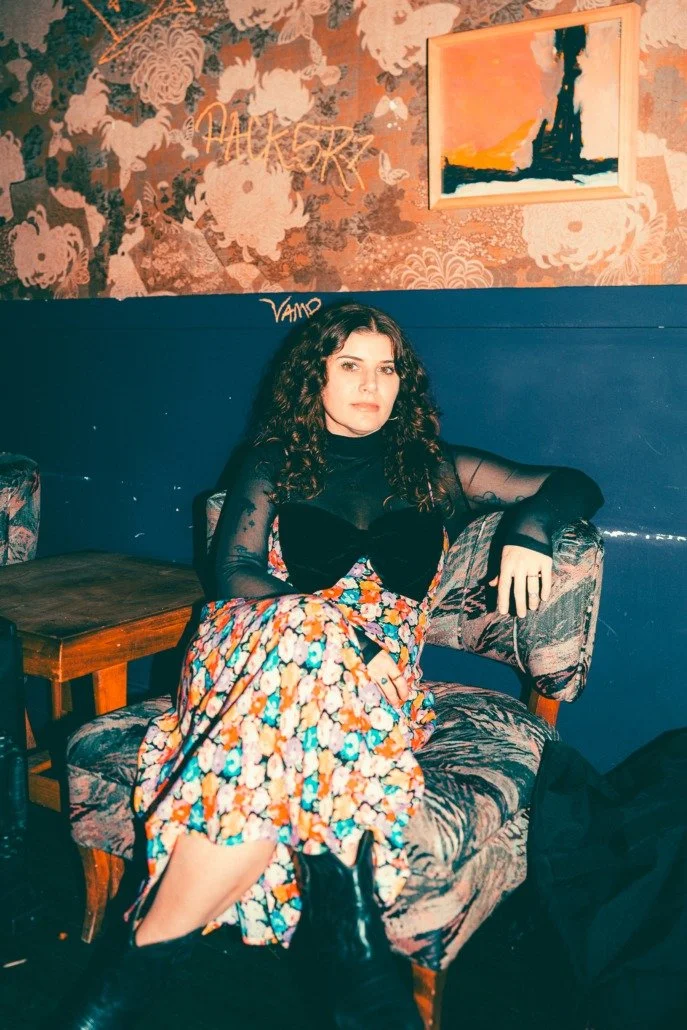 A woman with curly dark hair sitting on a vintage patterned armchair in a dimly lit room with floral wallpaper and abstract art on the wall.