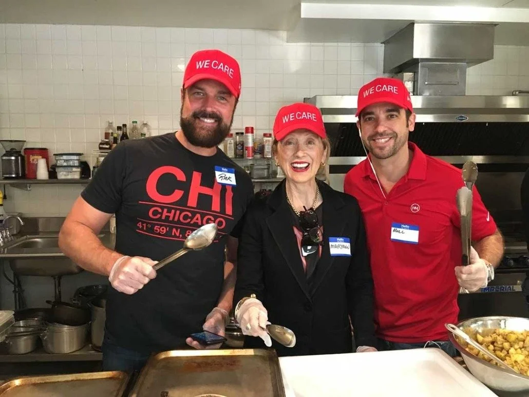 Three people, two men and one woman, standing in a kitchen, holding kitchen utensils and smiling. They are wearing red caps with white text that says "WE CARE." The woman is wearing a black jacket, and both men are in casual shirts—one black with a C