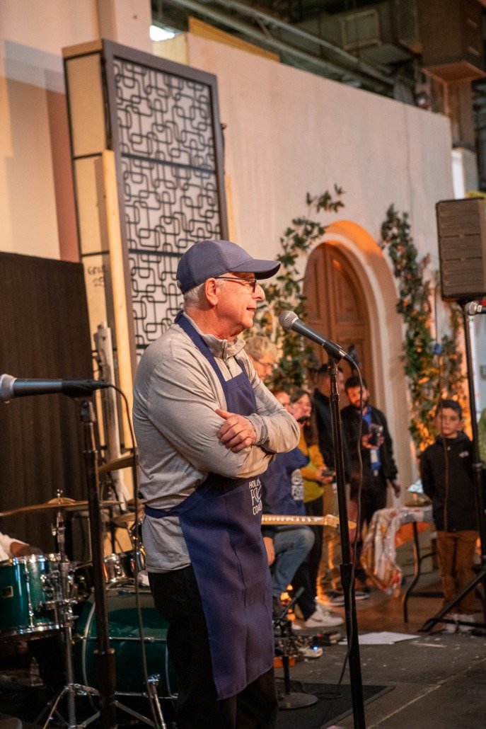 An elderly man wearing a blue baseball cap, glasses, gray long-sleeve shirt, and a blue apron standing with arms crossed near a microphone, in front of a group of people at an indoor event with musical instruments.