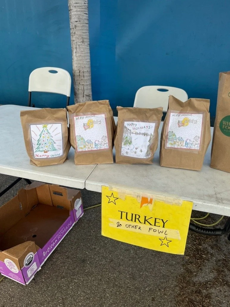 Table with four paper bags with holiday drawings and messages. Sign on the table reading "Turkey & Other Fowl" for a holiday donation or sale.