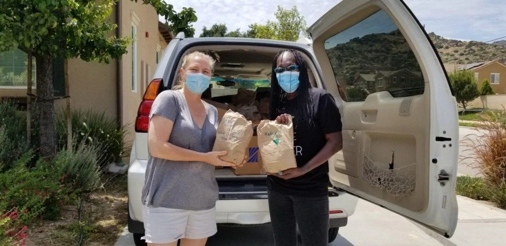 Two women wearing masks and casual clothes are standing next to an open SUV trunk, holding paper bags, in a suburban neighborhood on a sunny day.