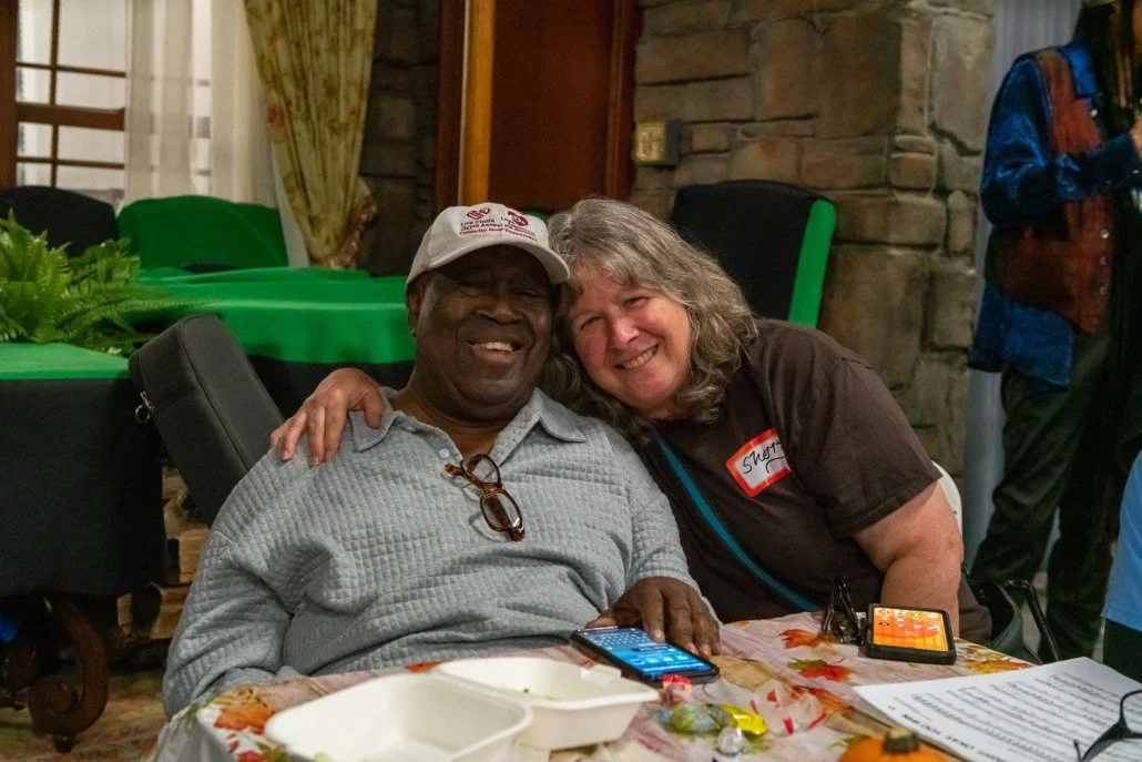 Two women happily sitting at a table, one with a gray shirt and a baseball cap, the other with a dark shirt and shoulder-length hair. They are smiling and leaning close together with one woman's arm around the other's shoulders.