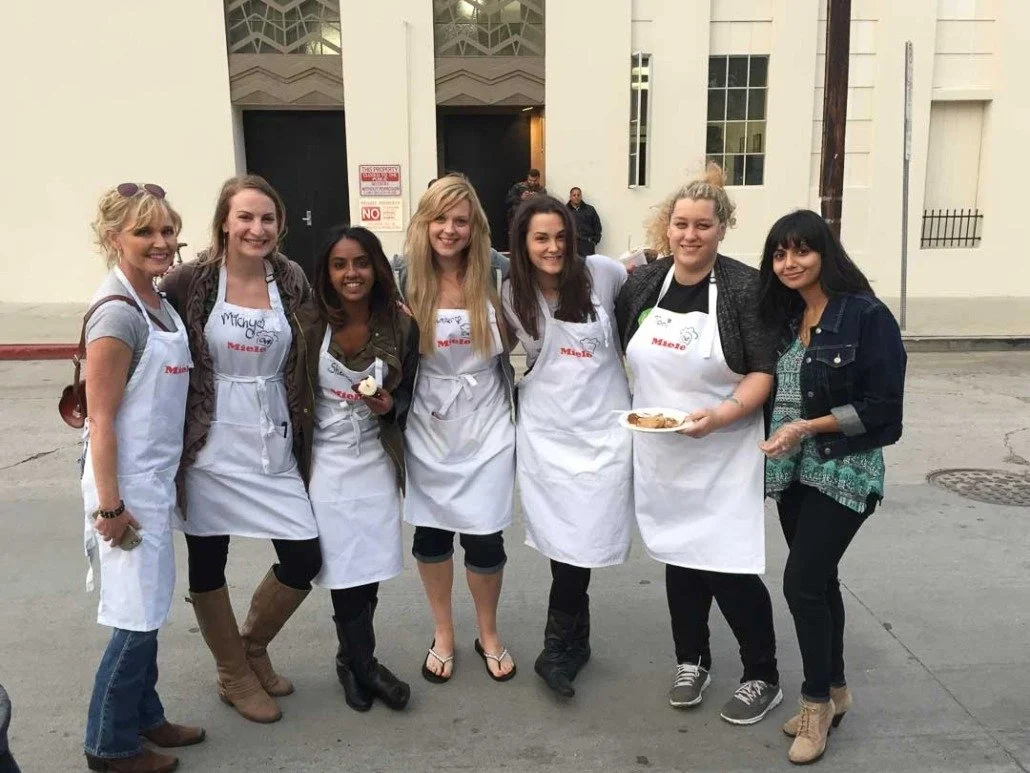 Group of seven women standing on a sidewalk wearing aprons, smiling, in front of a beige building, some holding plates of food.