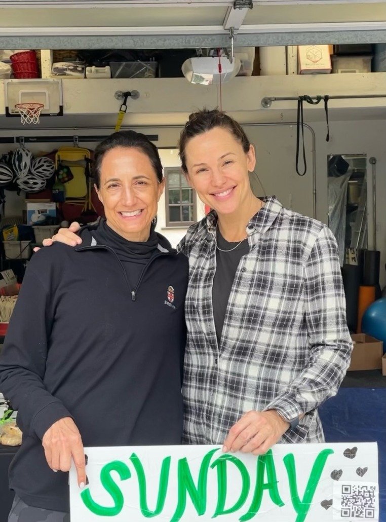 Two women smiling and holding a sign that reads 'SUNDAY' in a garage with sports gear and storage shelves in the background.