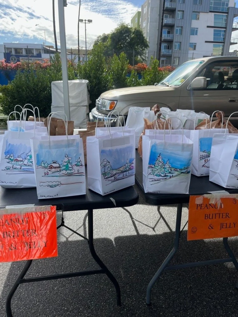 Table with paper bags labeled as peanut butter and jelly, set outdoors with a car and apartment buildings in background.