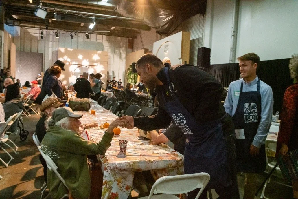 A man in a black jacket and apron shaking hands with an elderly woman wearing a green hoodie and cap seated at a long table during a community event.