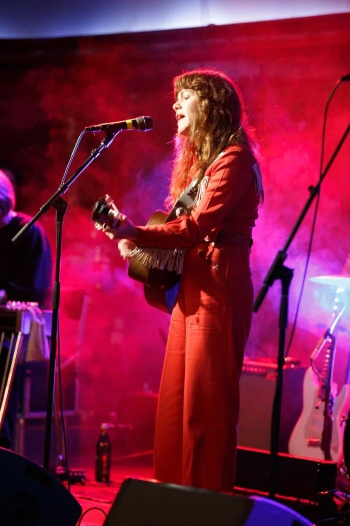 A woman with curly brown hair in a red outfit is singing and playing guitar on stage, with colorful stage lighting.