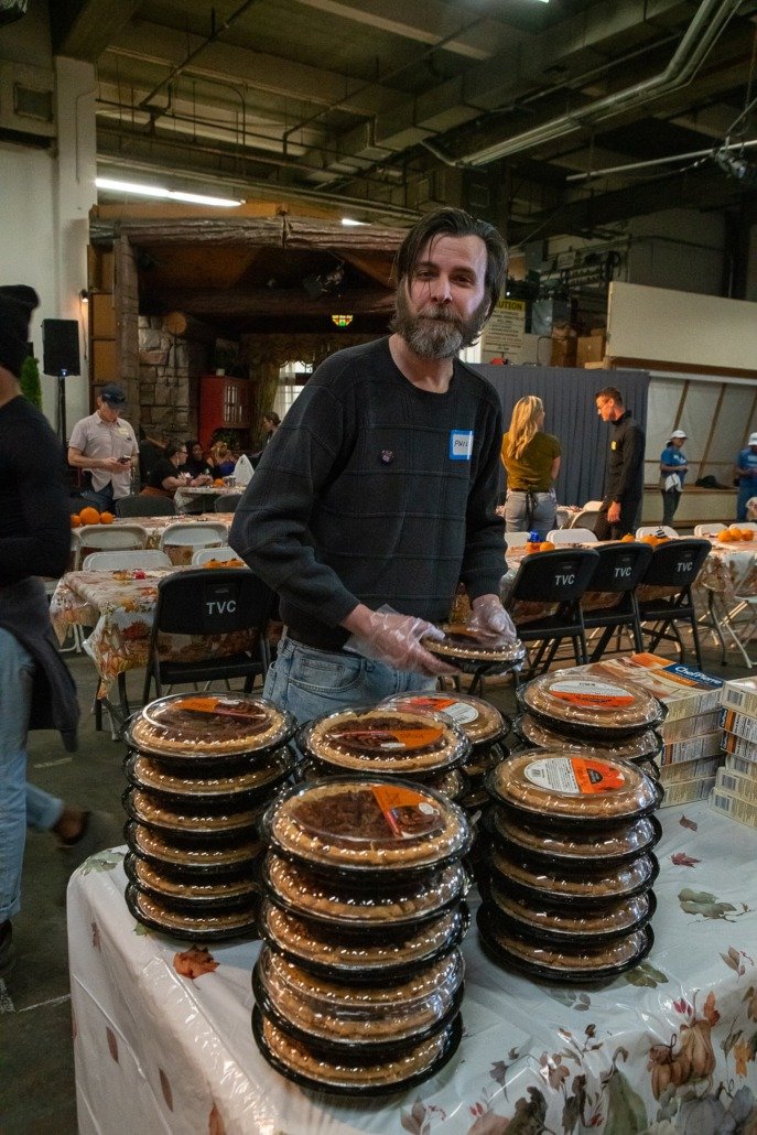 A man with a beard and dark hair, wearing a black sweater and gloves, stands behind a table stacked with packaged pies at an indoor event. The background shows other people, tables, and chairs, indicating a community gathering or market.