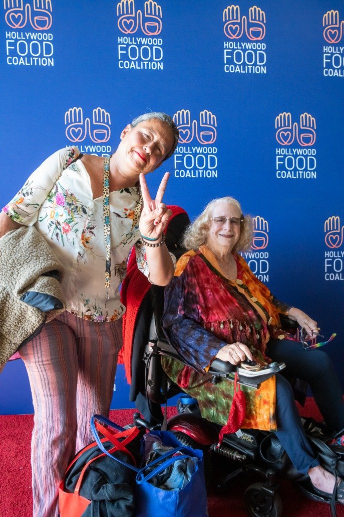 Two women, one standing and one seated in a wheelchair, posing in front of a blue backdrop with the Hollywood Food Coalition logo. The standing woman is making a peace sign and smiling.