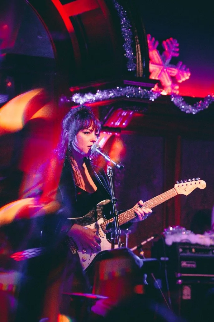 A female musician performs with an electric guitar and microphone on stage, decorated with Christmas ornaments and tinsel, under colorful stage lighting.