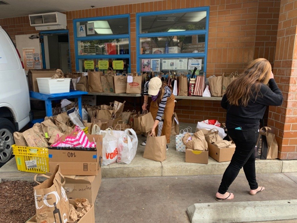 Two women are sorting through and organizing paper grocery bags and boxes outside a store or food bank. One woman is wearing a striped shirt and a beige hat, while the other has long brown hair and is dressed in black. Several brown paper bags and ca