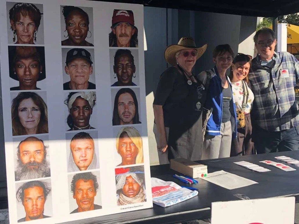 Group of people standing next to a display board with portraits of diverse individuals, outdoors with sunlight.