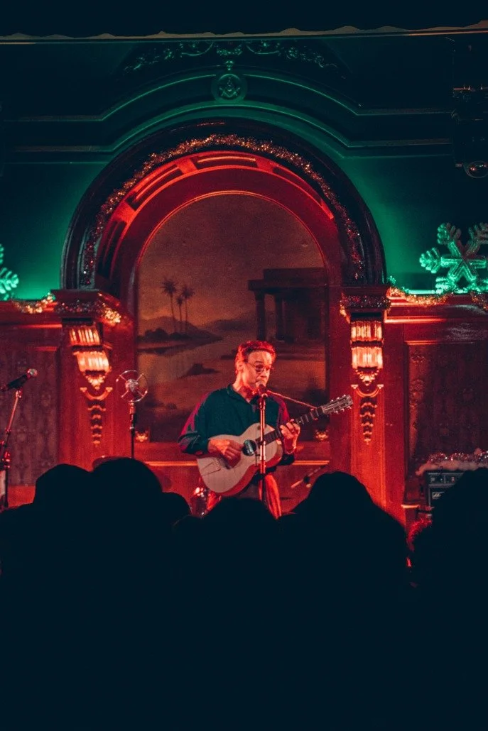 Person playing guitar and singing on stage with holiday decorations, including snowflake lights, in a dimly lit venue filled with audience.