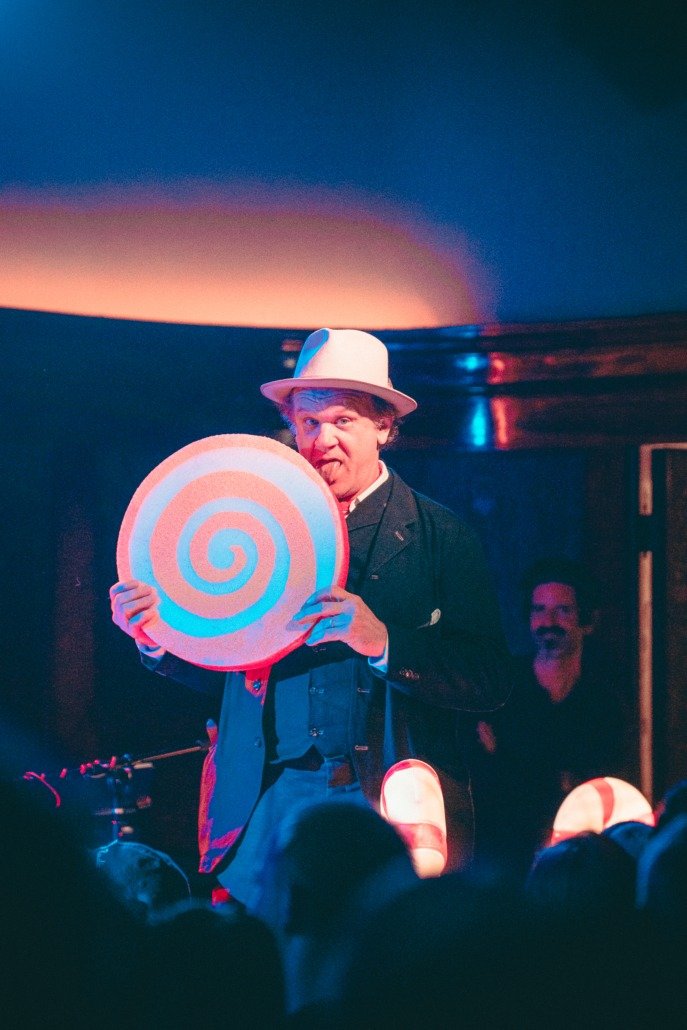 A person with a humorous expression holding a large circular object with a spiral pattern in front of a stage.