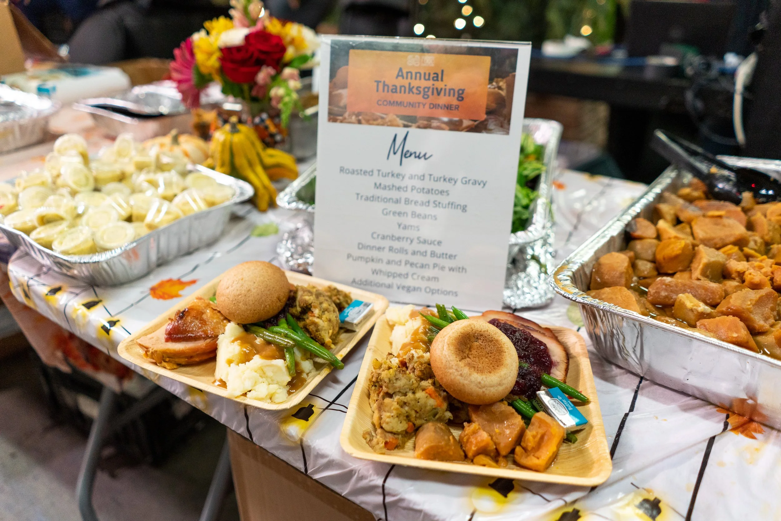 Food display at a community Thanksgiving dinner with trays of mashed potatoes, green beans, turkey, and various side dishes, along with plates of traditional Thanksgiving foods, a floral centerpiece, and a menu sign