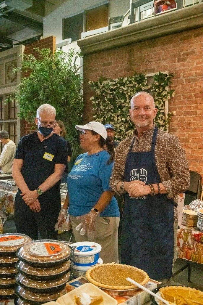 Group of three people at a food donation event, standing behind tables with pies and desserts, with a brick wall and green plants in the background.