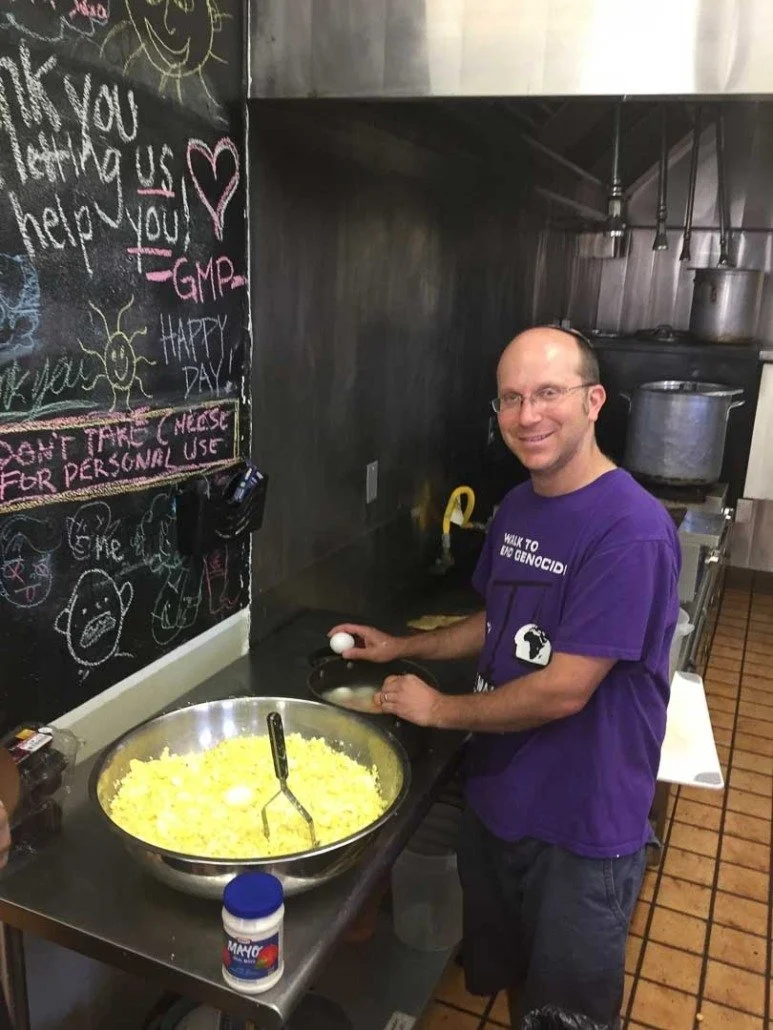 A man wearing glasses and a purple shirt that says 'Walk to End Genocide' is cooking scrambled eggs in a kitchen. He is holding an egg in his right hand. In front of him is a large mixing bowl of yellow scrambled eggs, with a spatula inside. The kitc