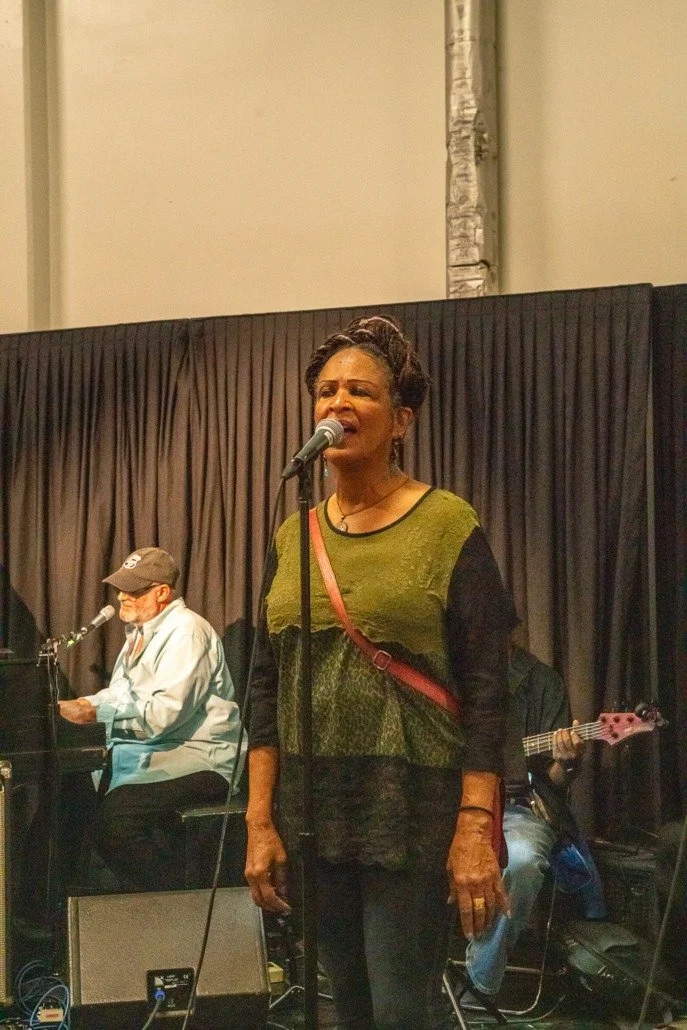 A woman singing into a microphone with a man playing piano and another person playing guitar in the background on stage with a black curtain.