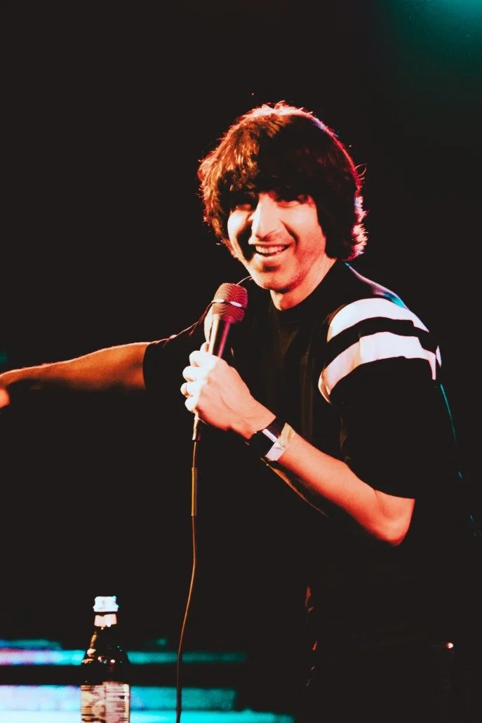A young man with dark, shaggy hair is holding a microphone and smiling on stage, wearing a black shirt with white stripes on the sleeves, with a dark background and stage lighting.