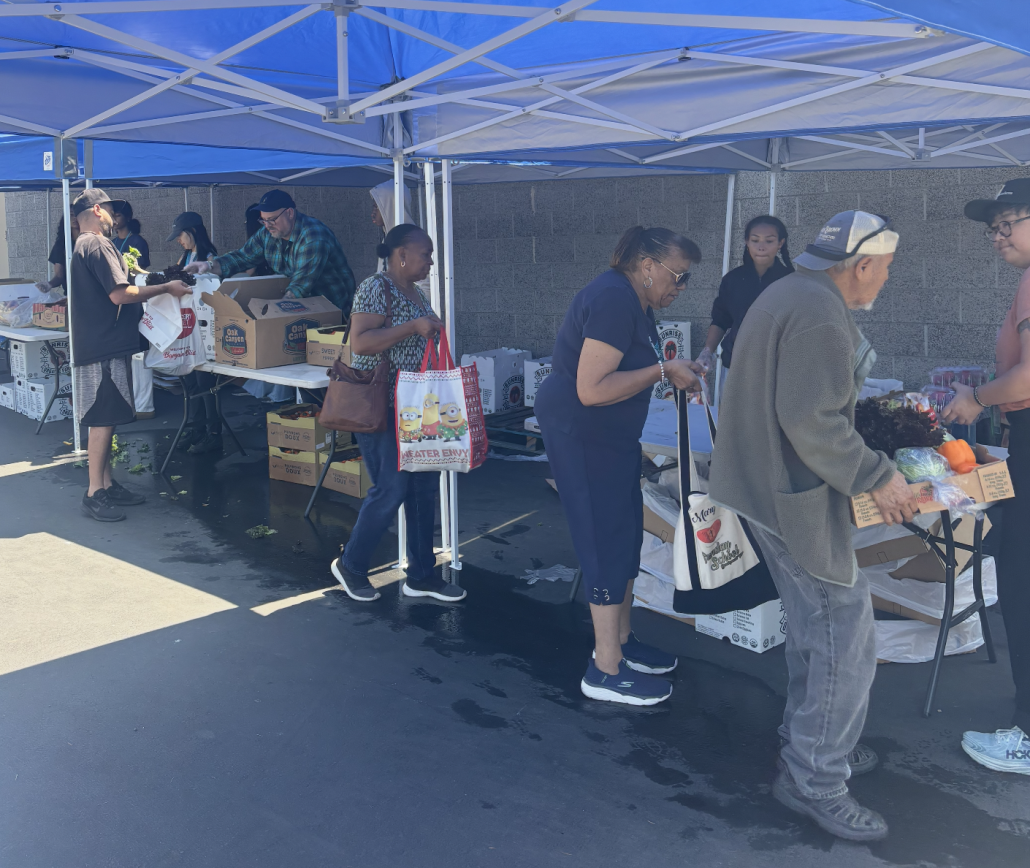 People shopping at a market stall under blue tents, buying and selling various goods like fruits and vegetables.