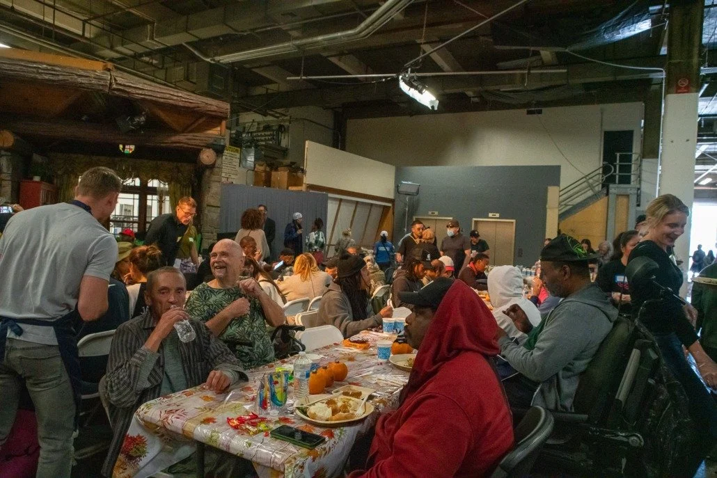 A large gathering of people sitting at long tables in an indoor space, possibly a community event or meal service, with some people serving and others eating or socializing.