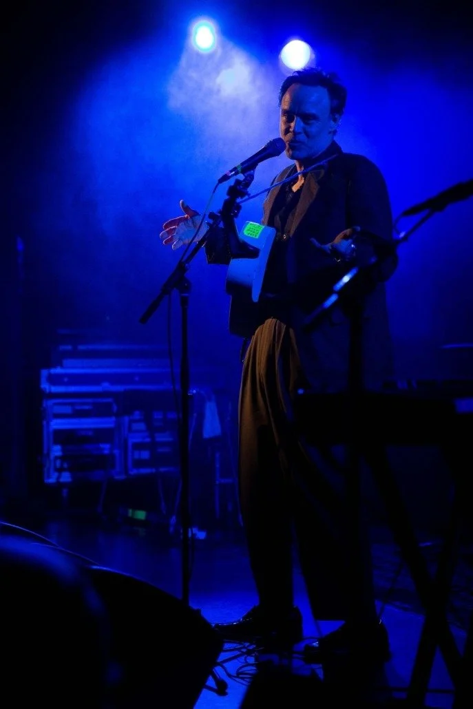 A man performing on stage with microphone, dressed in dark clothing, illuminated by blue stage lights, with musical equipment in the background.